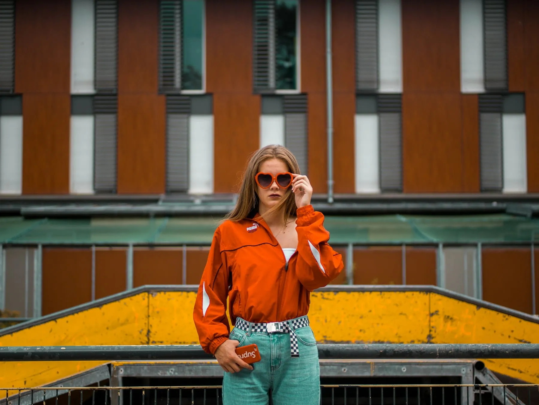 A person in orange sunglasses and a bright jacket posing against yellow city architecture