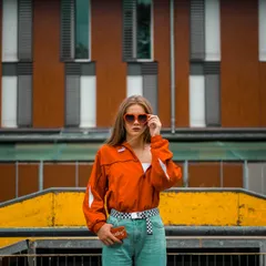 A person in orange sunglasses and a bright jacket posing against yellow city architecture